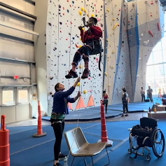 CJ Rose guides a person elevated by ropes & a swing in a rock climbing indoor space