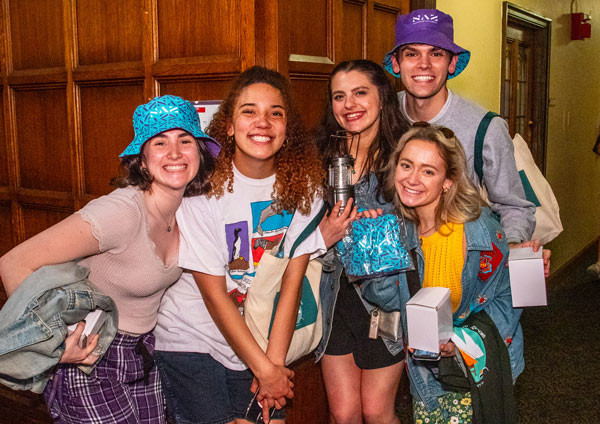 5 smiling students pose, wearing Naz hats and carrying merch, at Midnight Breakfast on campus 