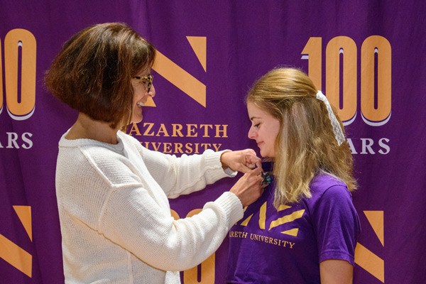 An adult attaches a legacy pin to the shirt of a relative who's a Naz student, by a Naz backdrop