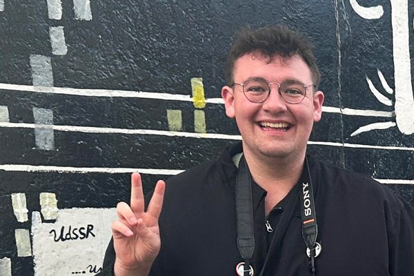 Matt Roberson makes a peace sign, has camera around neck, by mural in Berlin