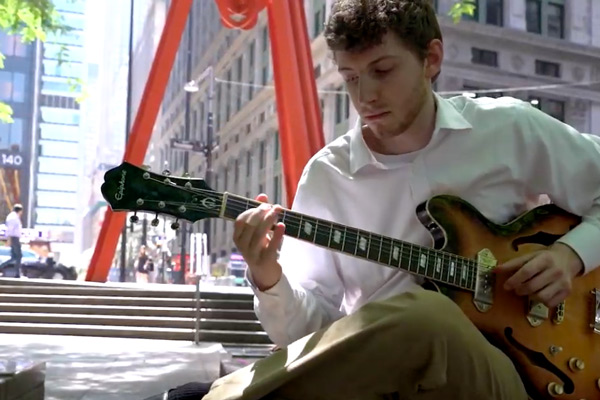 Sean Saville sits with guitar with New York City skyscrapers behind him