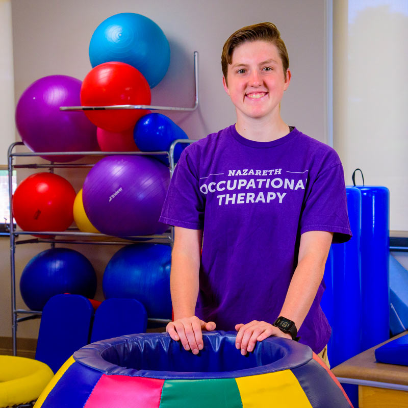 Megan Moyer poses, surrounded by colorful large balls and padded equipment for occupational therapy.