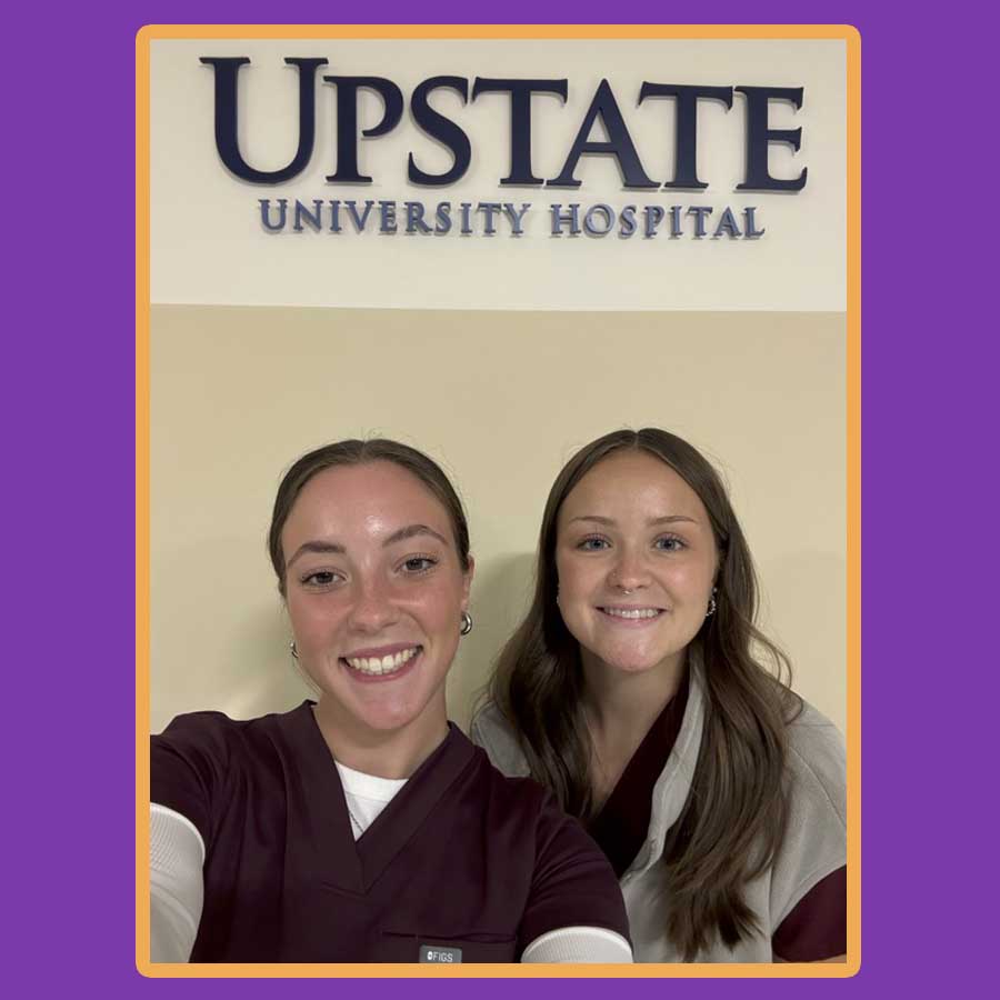 Selfie photo of two Nazareth nursing graduates posing in front of an Upstate University Hospital sign on the wall in the hospital. 