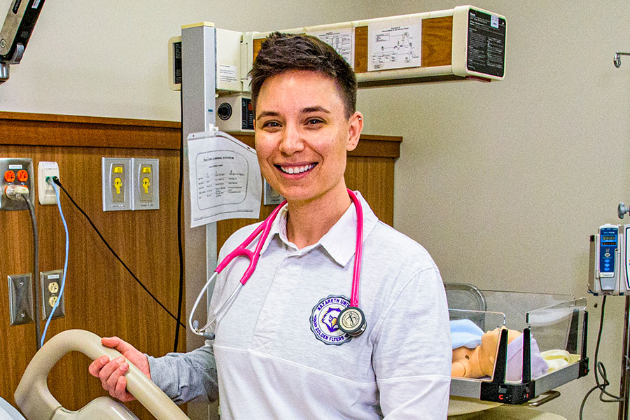 student smiling next to a hospital bed in the nursing sim lab