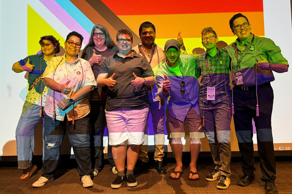 People pose with an inclusive pride flag of yellow, pink, light blue, brown, & primary colors