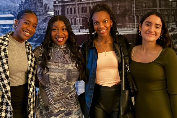 Four women in dresses, side by side, pose, smiling for the camera.