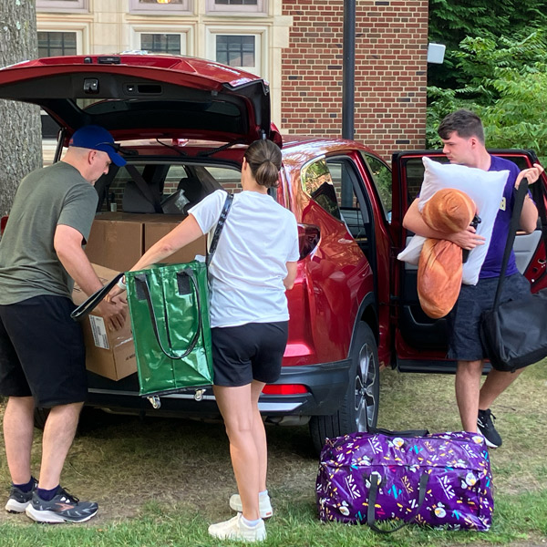 A family carries boxes, bags, and pillows from a car during move-in.