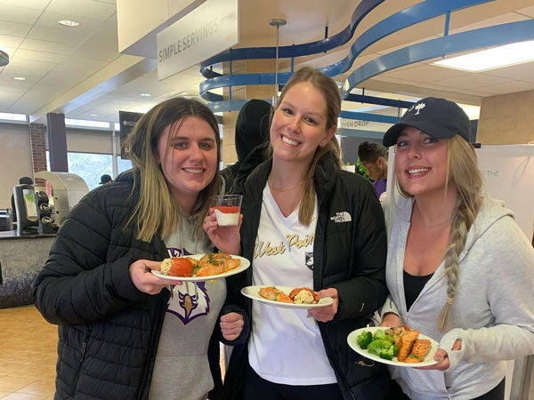 Three students in the Dining Commons hold plates of food and smile; sign says Simple Servings