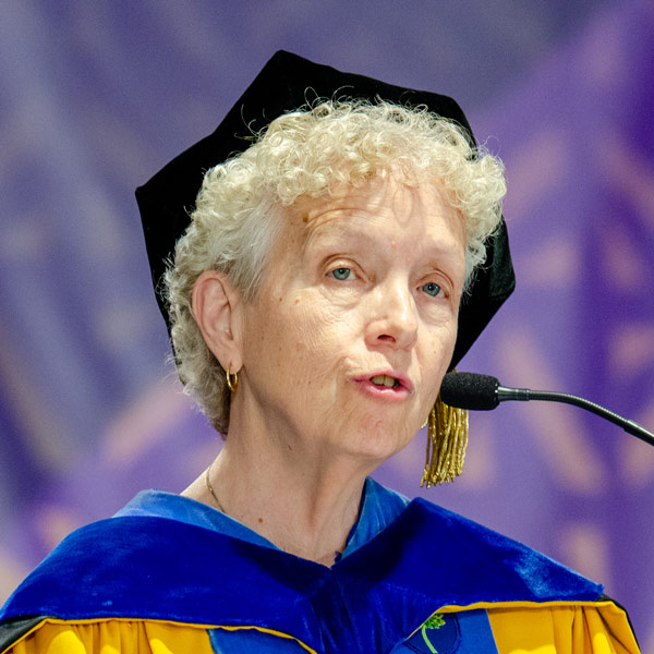 Mary Lou Mitchell wearing a commencement cap and robes speaks at podium at Commencement