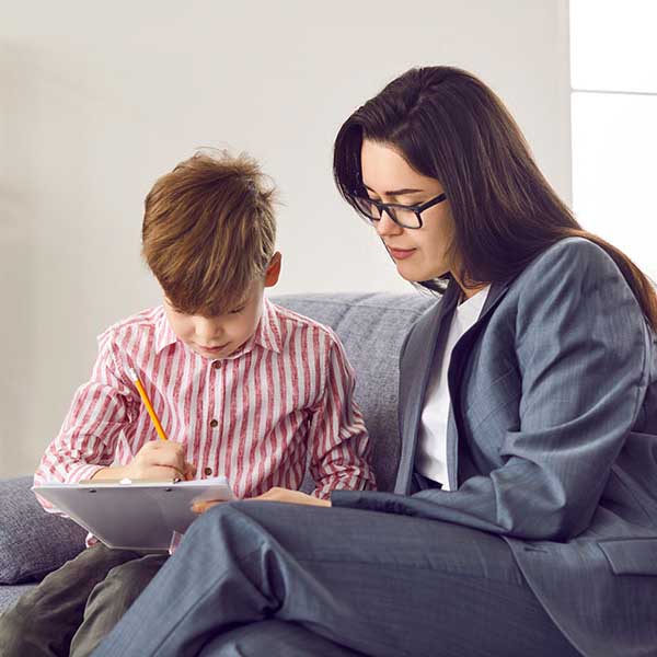 Female psychologist sits next to a small male child who is a client as he draws on a paper.