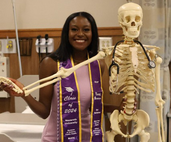 Bev Sainteme in Young Scholars, nursing graduation sash poses with skeleton, nursing simulation lab