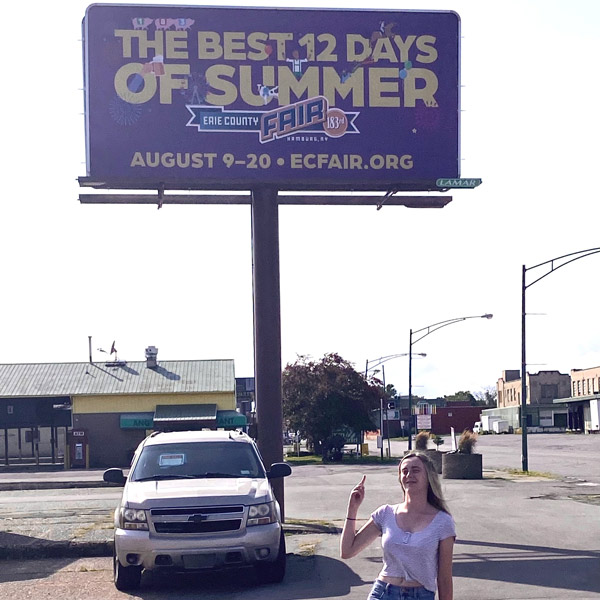 Annika poses on the ground below an Erie County Fair billboard she designed.