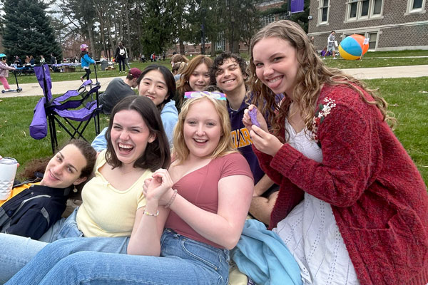 Seven students lean against each other in a clump on a campus lawn, smiling and laughing.