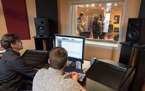 Students use digital equipment in music studio, seeing musicians through window 