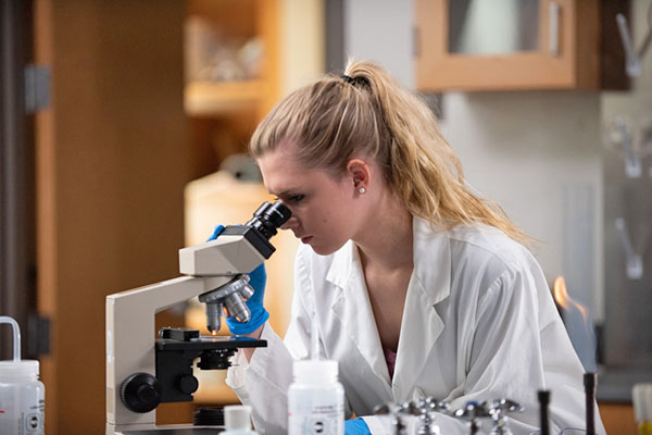 student in lab coat looking through a microscope