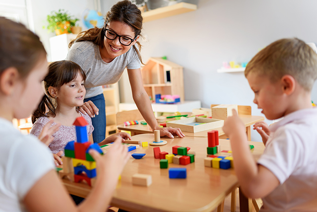 three children at a table building with colorful blocks with adult watching