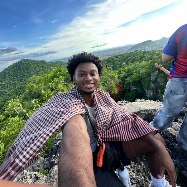 Selfie:Cloth tied around shoulders flies out behind Denim Powers. He's above treetops on a mountain.