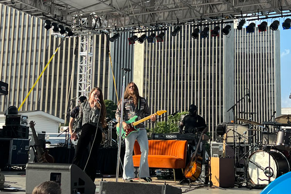 A band performs on an outdoor stage with skyscrapers in the background and a couch onstage