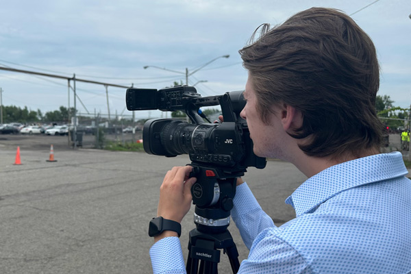 Stevie Skvarek looks through viewfinder of a videocamera on a tripod, traffic cones in distance