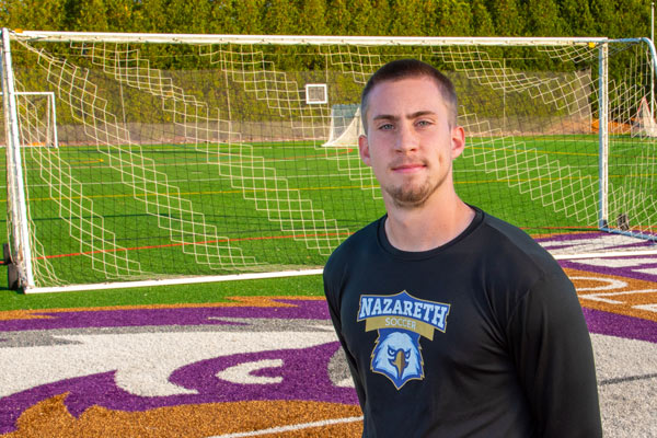 Alex Gent poses on soccer field, wearing a Nazareth University Golden Flyer athletics logo on hshirt