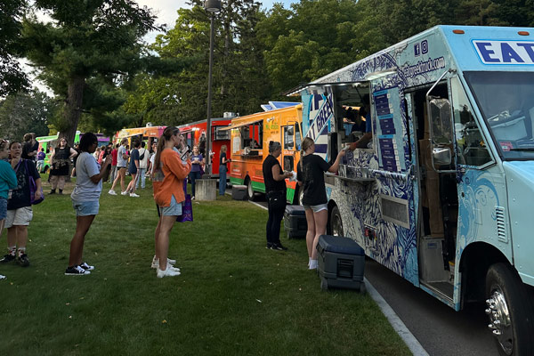 People sit at purple-topped tables near a line of parked food trucks